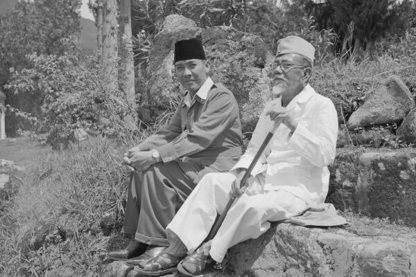 Haji Agus Salim duduk berdampingan dengan Presiden Soekarno di luar ruangan, mengenakan pakaian putih dan memegang tongkat.