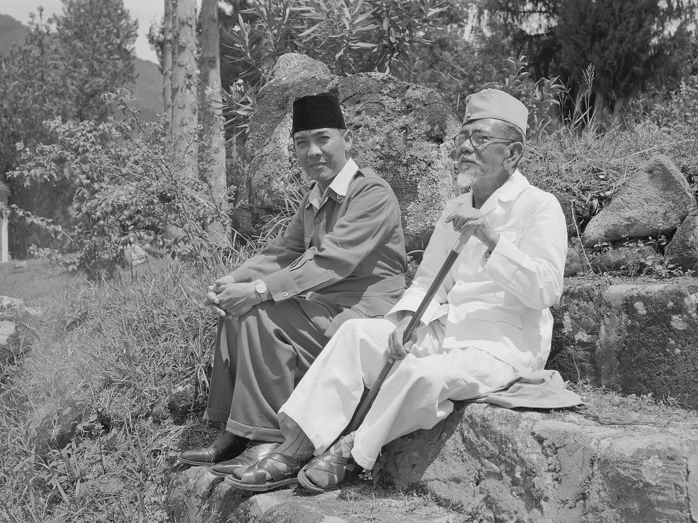 Haji Agus Salim duduk berdampingan dengan Presiden Soekarno di luar ruangan, mengenakan pakaian putih dan memegang tongkat.