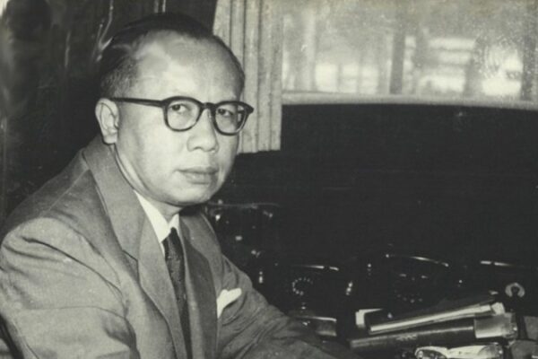 Ir. H. Djuanda Kartawidjaja sedang duduk di meja kerjanya, mengenakan jas dan kacamata