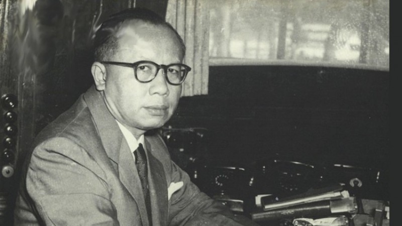 Ir. H. Djuanda Kartawidjaja sedang duduk di meja kerjanya, mengenakan jas dan kacamata