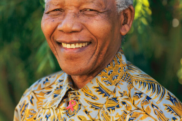 Nelson Mandela tersenyum mengenakan kemeja bermotif, latar belakang pepohonan