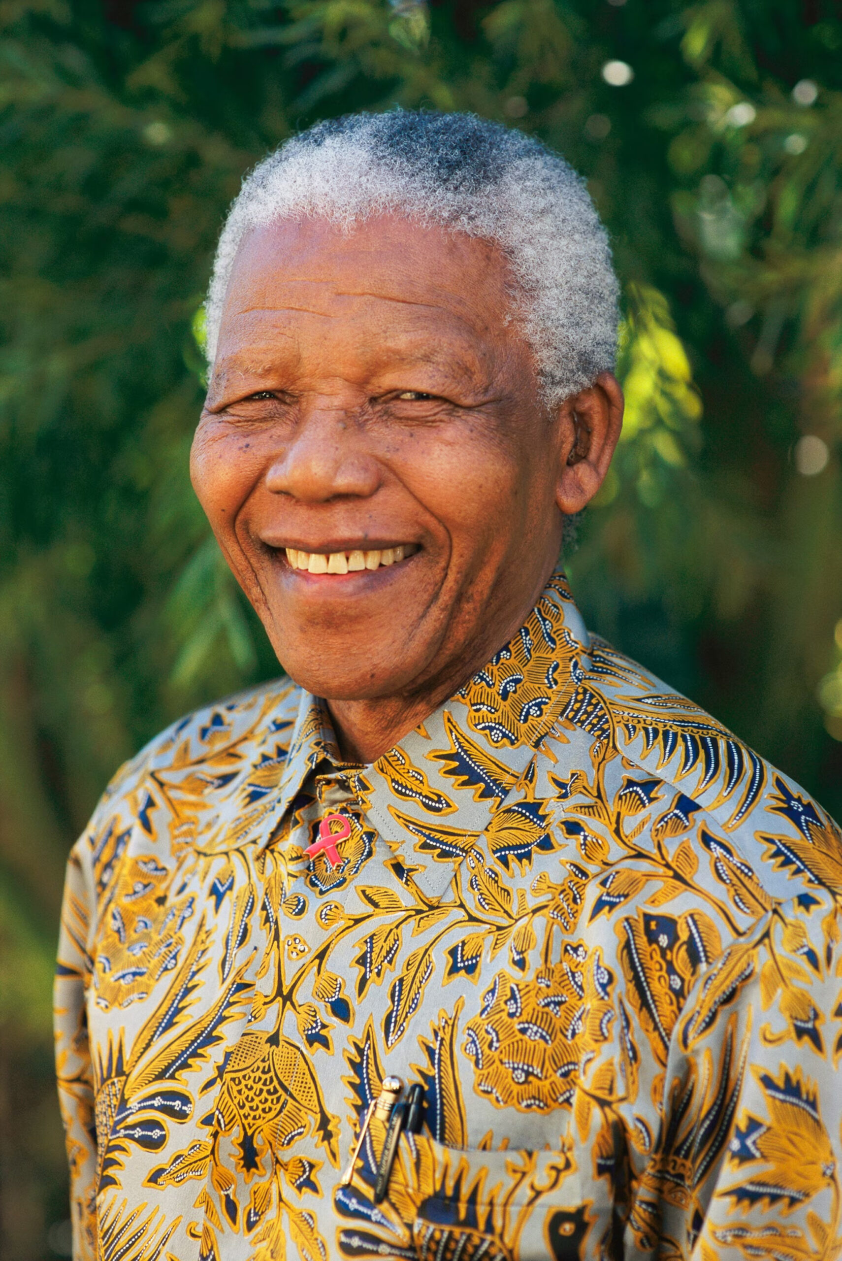 Nelson Mandela tersenyum mengenakan kemeja bermotif, latar belakang pepohonan