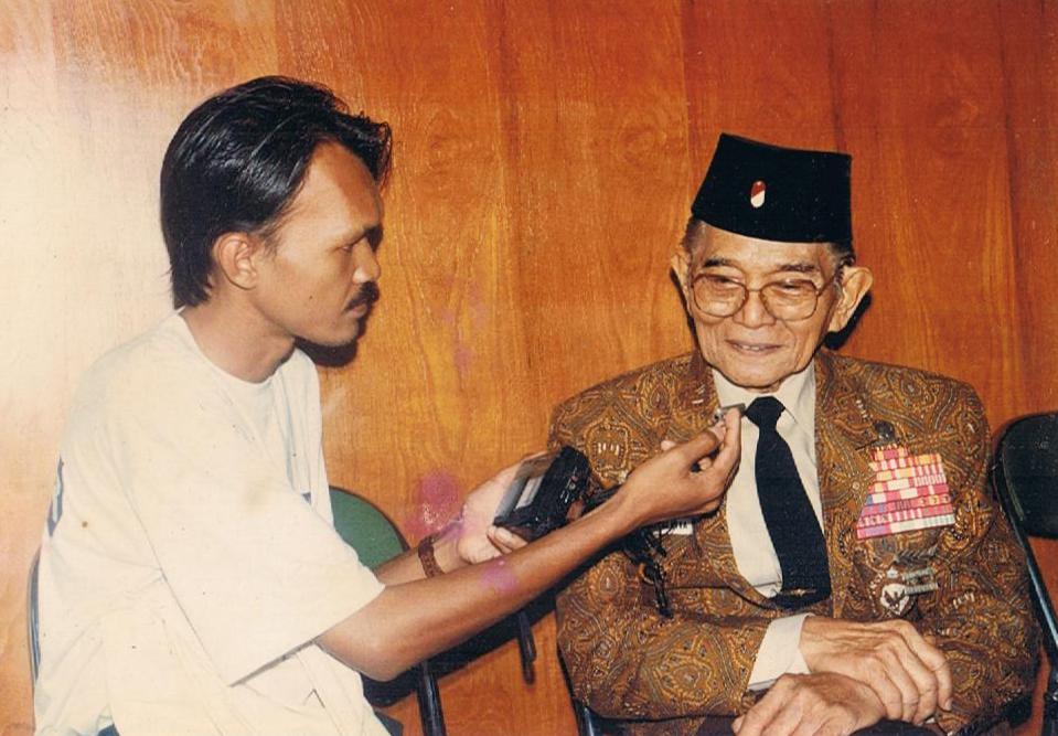 Andi Mattalatta diwawancarai wartawan, mengenakan jas batik dan peci.